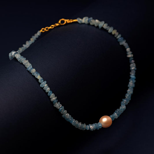 "Luxora Necklace with ocean blue natural stone chips and pearl centerpiece in 18K PVD gold plated stainless steel clasp on dark cylindrical surface with navy background"