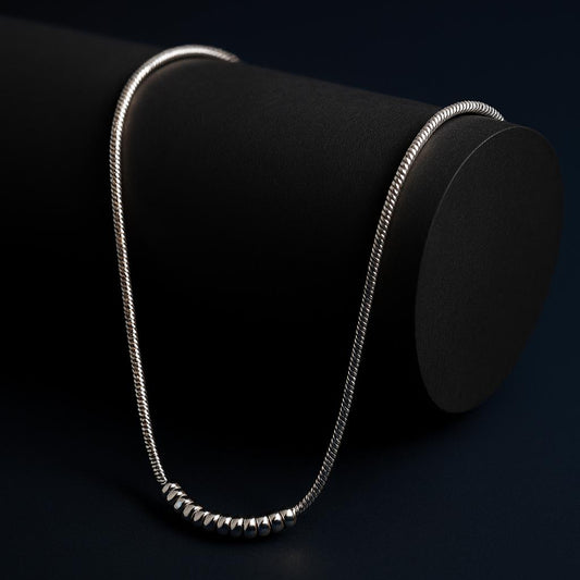 SILVER DAWN NECKLACE – Minimalist snake chain with spiral beads in 18K PVD silver plating displayed on black cylindrical surface