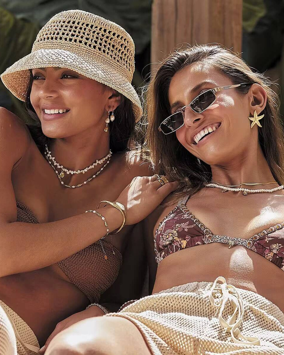 Two women in beach attire with hats and sunglasses, smiling outdoors.